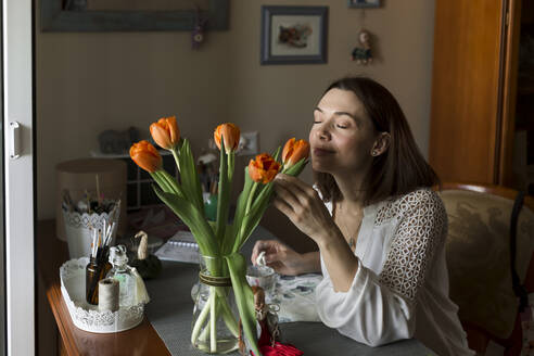 Smiling mature woman smelling tulips in vase on table at home - LLUF00522