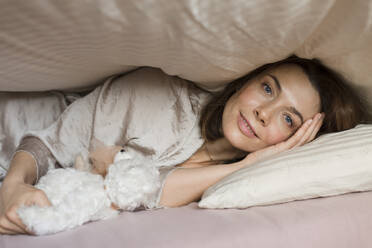 Smiling mature woman with teddy bear toy lying under blanket on bed at home - LLUF00525