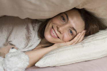 Happy mature woman with teddy bear toy lying under blanket on bed at home - LLUF00526