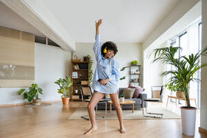 Young woman listening music through headphones dancing with hand raised in living room at home - OIPF01768