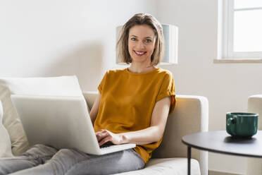 Smiling woman with laptop sitting on sofa in living room - DIGF17891