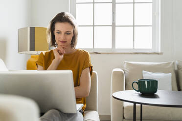 Smiling freelancer with hand on chin looking at laptop siting in living room - DIGF17896