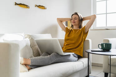 Smiling woman with hands behind head sitting on sofa in living room - DIGF17898