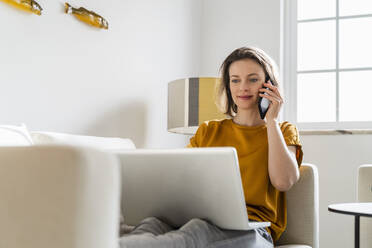 Smiling freelancer talking on mobile phone sitting with laptop on sofa in living room - DIGF17900