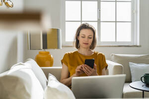 Freelancer using smart phone sitting with laptop on sofa at home - DIGF17902