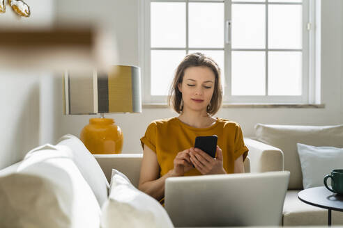 Freiberufler mit Smartphone und Laptop auf dem Sofa zu Hause - DIGF17902