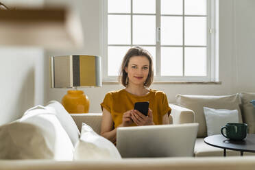 Smiling freelancer with smart phone and laptop sitting on sofa in living room - DIGF17903