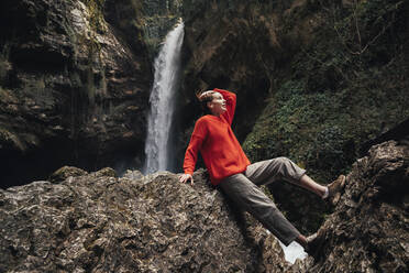 Junge Frau sitzt auf einem Felsen vor einem Wasserfall - OMIF00793