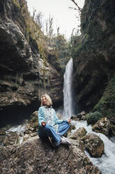 Ältere Frau sitzt auf einem Felsen vor einem Wasserfall - OMIF00794