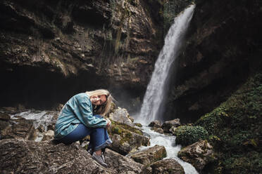 Glückliche reife Frau sitzt auf einem Felsen vor einem Wasserfall - OMIF00795