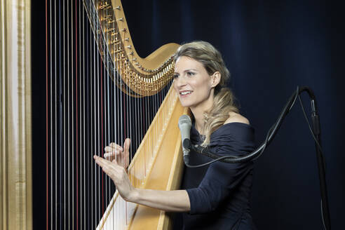 Smiling mature woman playing harp in front of black background - RNF01354