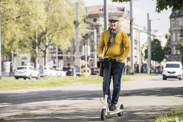 Smiling senior man riding electric push scooter on sidewalk in the city - UUF26013