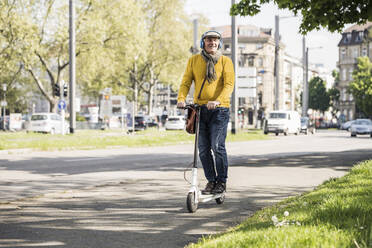 Smiling senior man wearing wireless headphones riding on electric push scooter - UUF26016