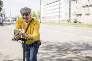 Lächelnder älterer Mann, der ein Mobiltelefon benutzt und sich auf einen elektrischen Motorroller stützt - UUF26017