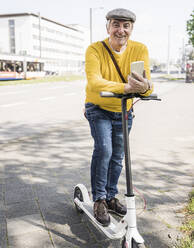 Smiling senior man with mobile phone and electric push scooter on sidewalk - UUF26018