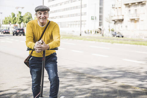 Glücklicher älterer Mann mit verschränkten Händen, der sich auf einen elektrischen Motorroller stützt - UUF26021