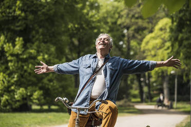 Carefree senior man with arms outstretched riding bicycle at park - UUF26023