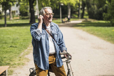 Smiling senior man wearing In-ear headphones with bicycle at park - UUF26024