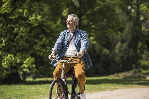 Lächelnder Mann mit Brille auf dem Fahrrad im Park an einem sonnigen Tag - UUF26027