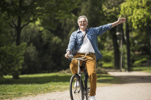 Glücklicher älterer Mann mit erhobenen Armen, der eine Fahrradtour im Park genießt - UUF26028