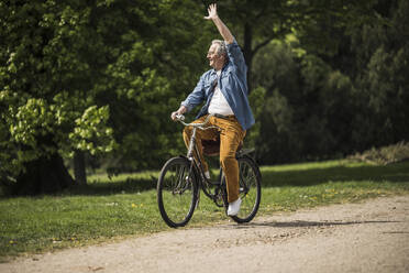 Smiling senior man riding bicycle waving hand at park on sunny day - UUF26029