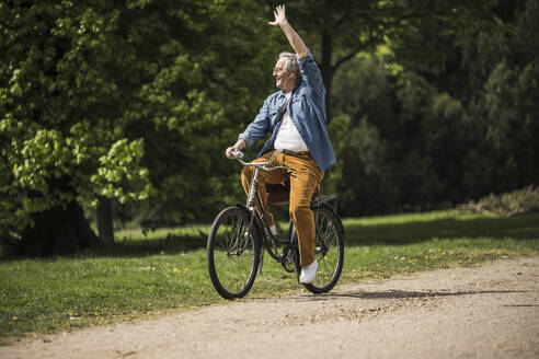 Lächelnder älterer Mann auf dem Fahrrad und winkende Hand im Park an einem sonnigen Tag - UUF26029