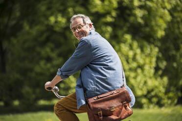 Happy senior man with crossbody bag riding bicycle at park - UUF26031
