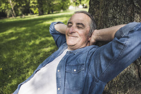 Lächelnder älterer Mann mit geschlossenen Augen, der sich im Park an einen Baumstamm lehnt - UUF26033