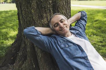 Smiling retired senior man with eyes closed leaning on tree trunk at park - UUF26034