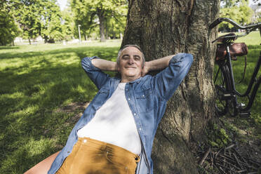 Smiling man with fully unbuttoned denim shirt leaning on tree trunk at park - UUF26036