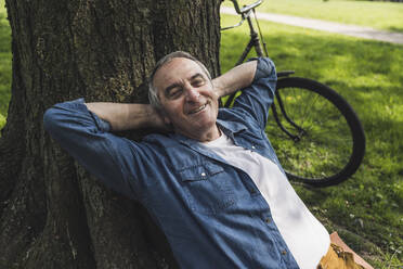 Happy senior man leaning on tree trunk by bicycle at park - UUF26037