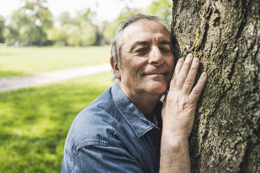 Lächelnder älterer Mann mit geschlossenen Augen umarmt Baum im Park - UUF26044