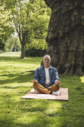 Senior man with eyes closed meditating on exercise mat at park - UUF26053