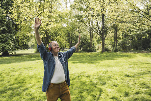 Happy senior man with arms raised enjoying music through wireless headphones at park - UUF26059