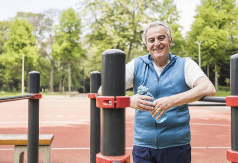Smiling senior man holding water bottle standing amidst gymnastics bar - UUF26063