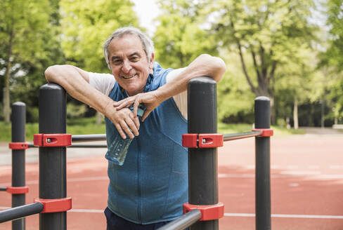 Smiling senior man holding water bottle standing amidst gymnastics bar at park - UUF26064