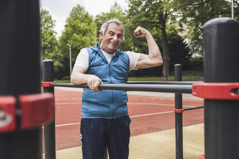 Happy senior man flexing muscles standing by gymnastics bar - UUF26072