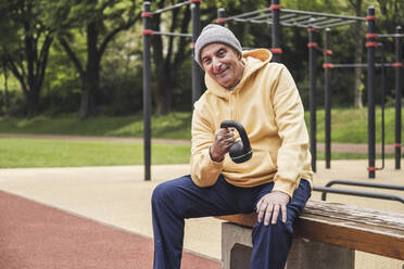 Glücklicher älterer Mann mit Kettlebell auf einer Bank im Park sitzend - UUF26075