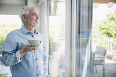 Glücklicher älterer Mann mit Kaffeetasse, der durch ein Fenster hinausschaut - UUF26093