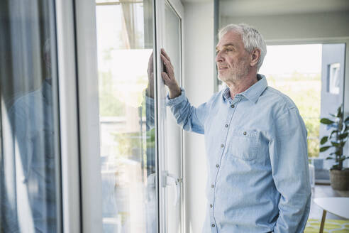 Lächelnder älterer Mann schaut durch das Fenster seines Hauses - UUF26095