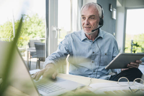 Senior businessman wearing headset working on laptop at home - UUF26101