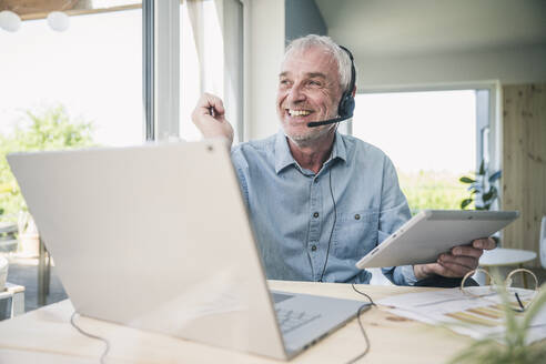 Glücklicher Freiberufler mit Headset, der zu Hause mit Tablet-PC und Laptop sitzt - UUF26106