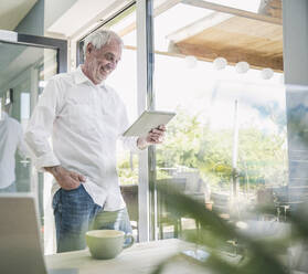 Happy senior man using tablet PC standing by glass window at home - UUF26110