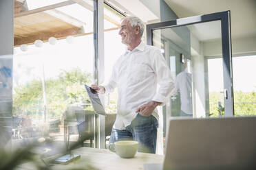 Smiling senior man with tablet PC standing glass window at home - UUF26111