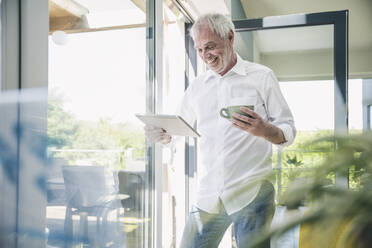 Happy senior man with tablet PC and coffee cup standing by glass window - UUF26112