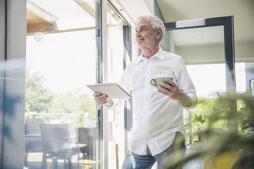 Lächelnder älterer Mann mit Tablet-PC und Kaffeetasse vor einem Glasfenster - UUF26113