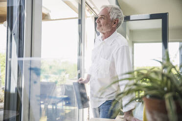Lächelnder älterer Mann mit Tablet-PC, der sich auf einen Tisch stützt, um zu Hause zu träumen - UUF26114