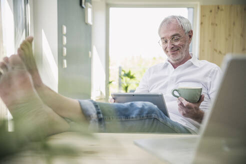 Lächelnder älterer Mann mit Kaffeetasse und Tablet-PC zu Hause sitzend - UUF26119