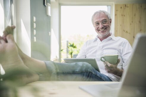 Glücklicher älterer Mann mit Kaffeetasse und Tablet-PC zu Hause sitzend - UUF26120