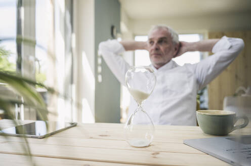 Hourglass on table with senior man sitting in background at home - UUF26121